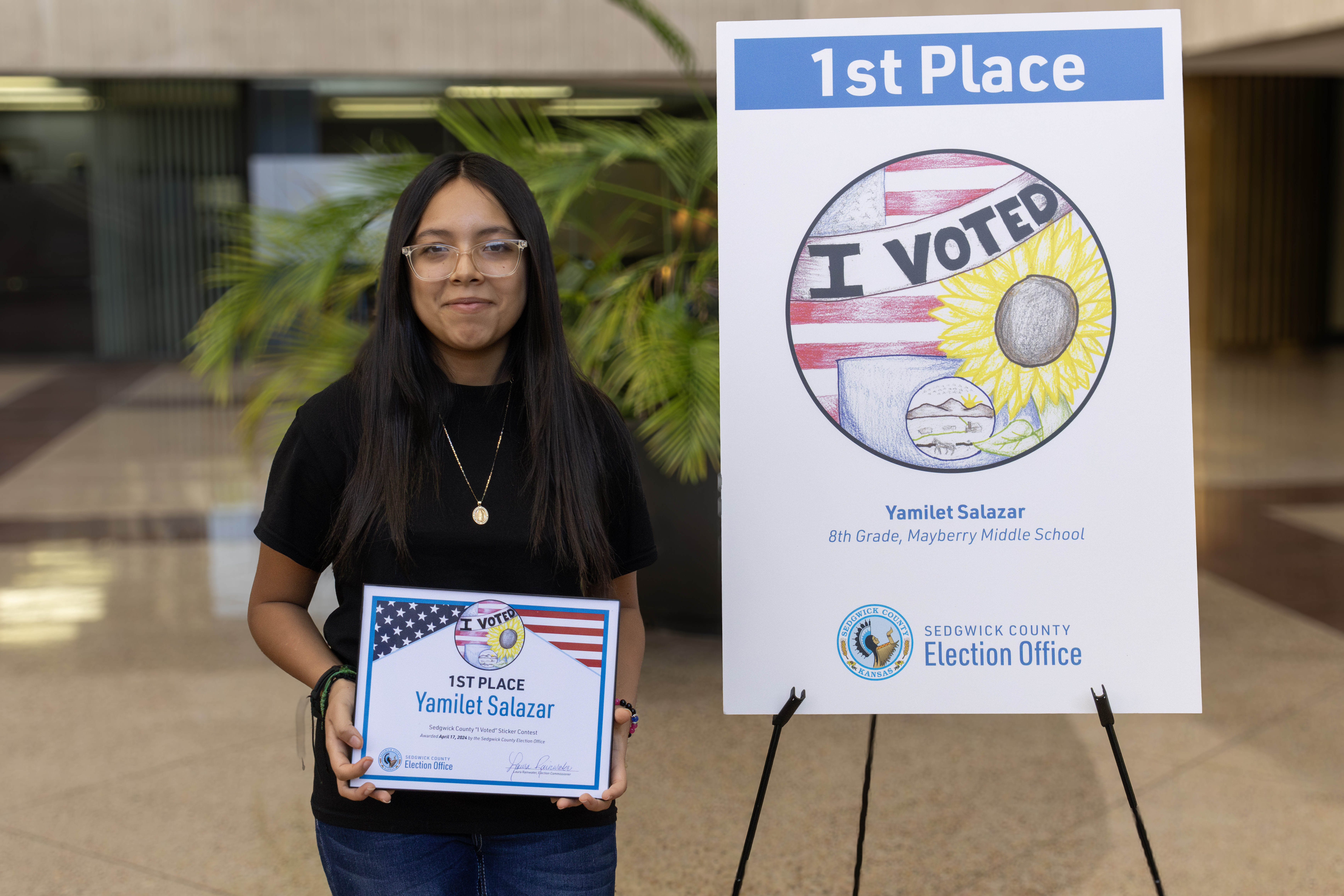1st place winner standing next to a tripod displaying her sticker. Her sticker is the american flag, a sunflower and the Kansas flag with a banner of I Voted.