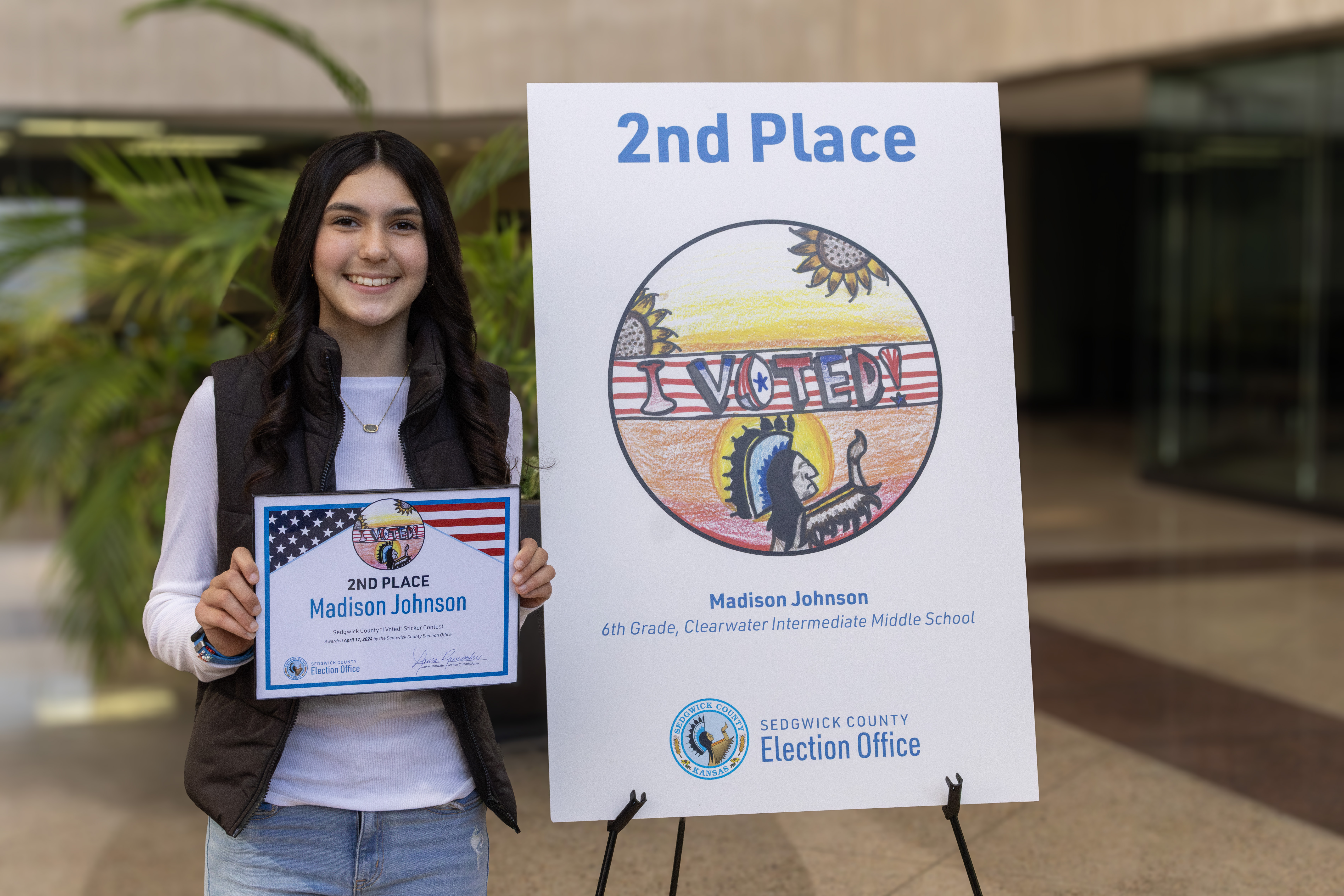 2nd place tie winner standing next to her sticker on a tripod. Her sticker is a drawn image of the keeper of the plains with a sun and sunflower background. A banner text I voted in the foreground.
