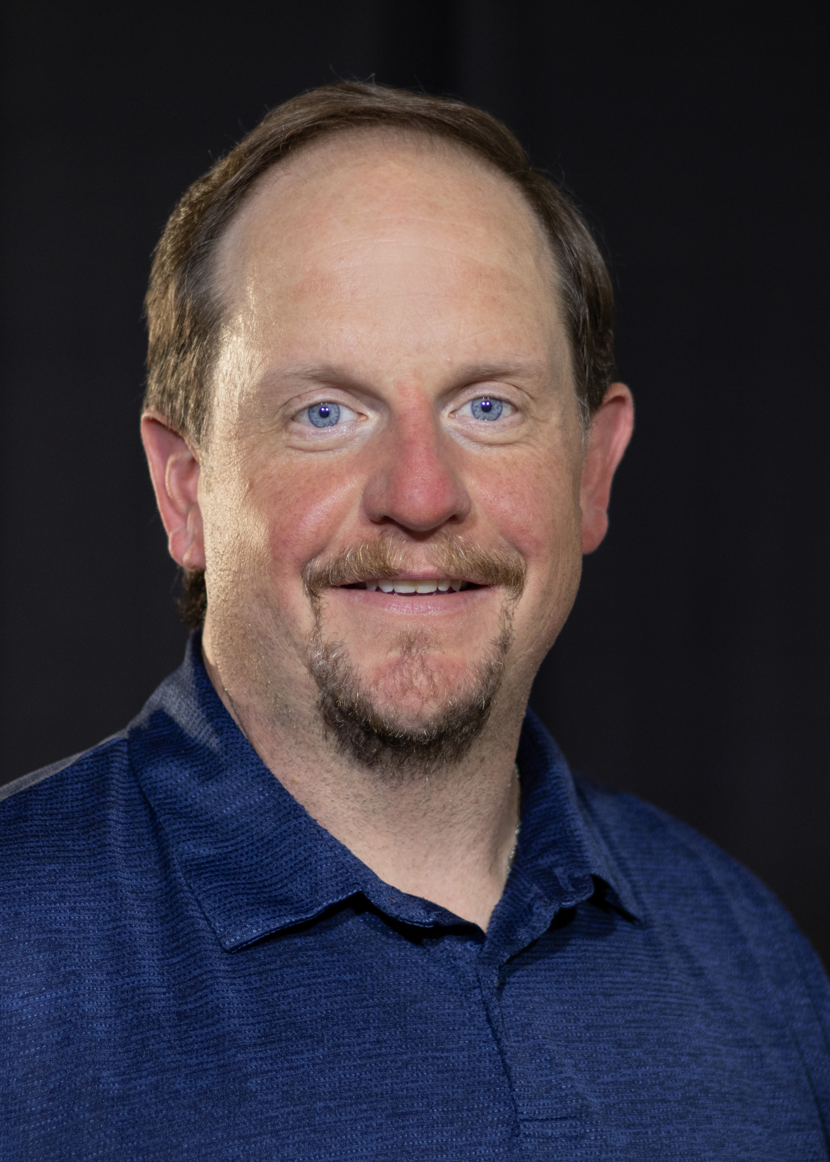 A picture of the Executive Director who is a man with short brown hair and goatee, wearing a blue shirt.
